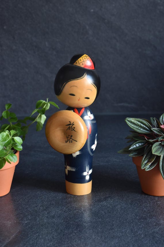 Miyashita Hajime kokeshi doll holding a round wooden hat inscribed with the piece title next to its sign