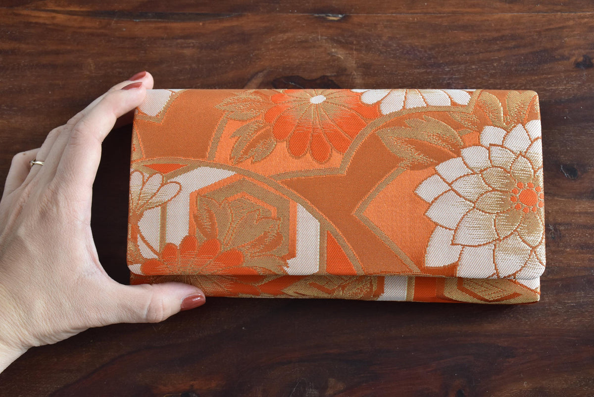 Saga Nishiki brocade clutch handbag shown next to a hand for size comparison on a dark wooden surface.