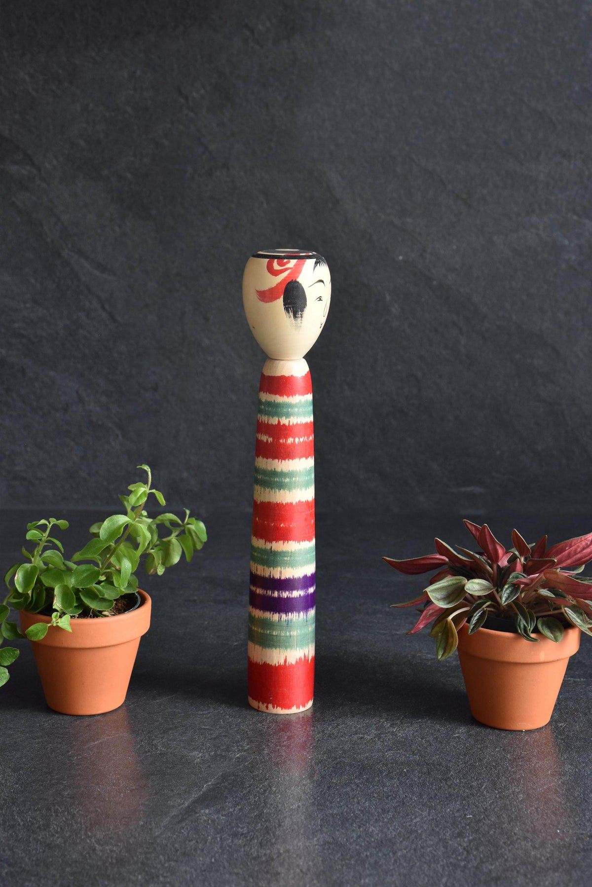 Tsuchiyu-Kei Kokeshi Doll Lathe-Spun Bands shown upright in full view with plants on both sides