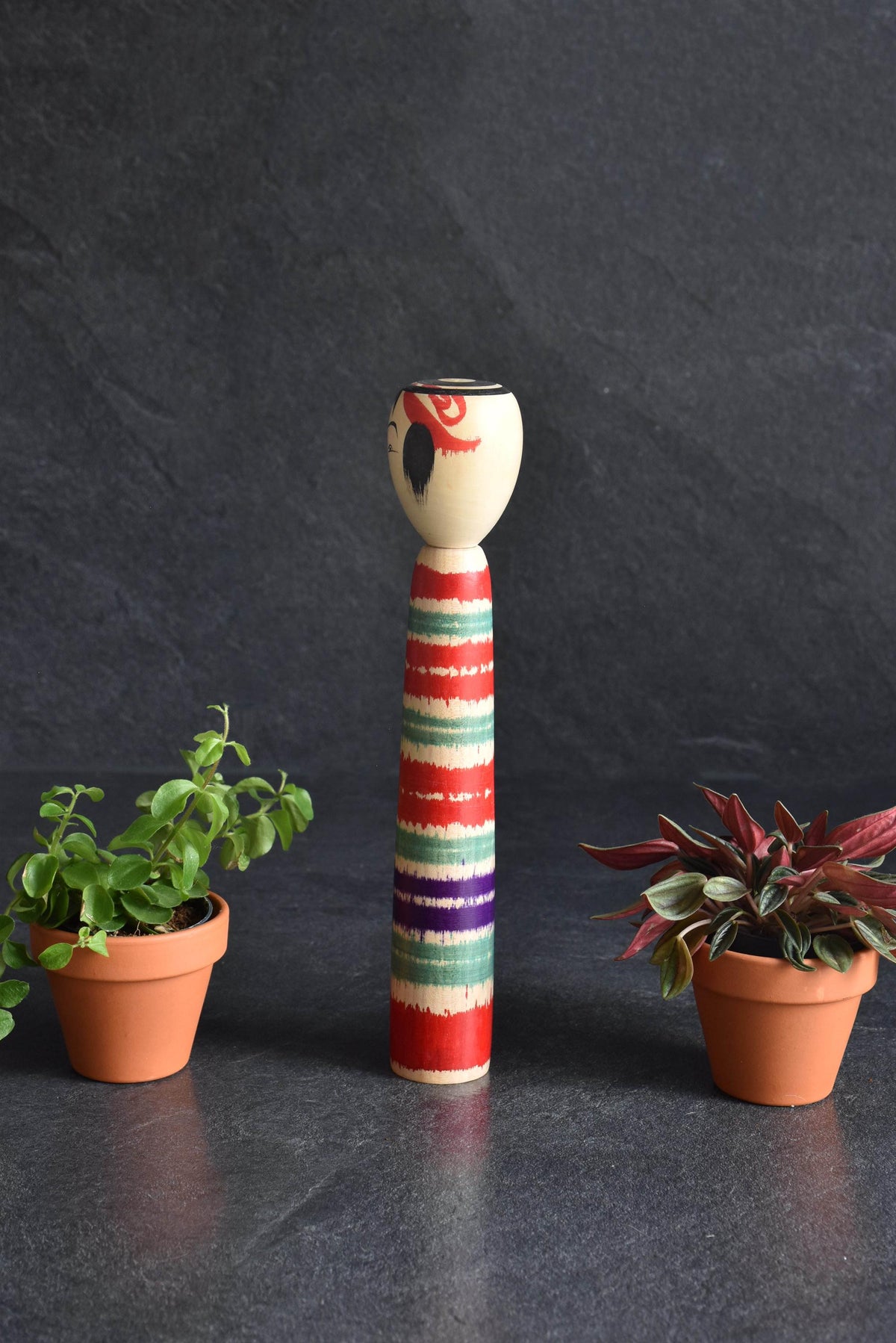 Tsuchiyu-Kei Kokeshi Doll Lathe-Spun Bands viewed from a slightly different angle between two small potted plants
