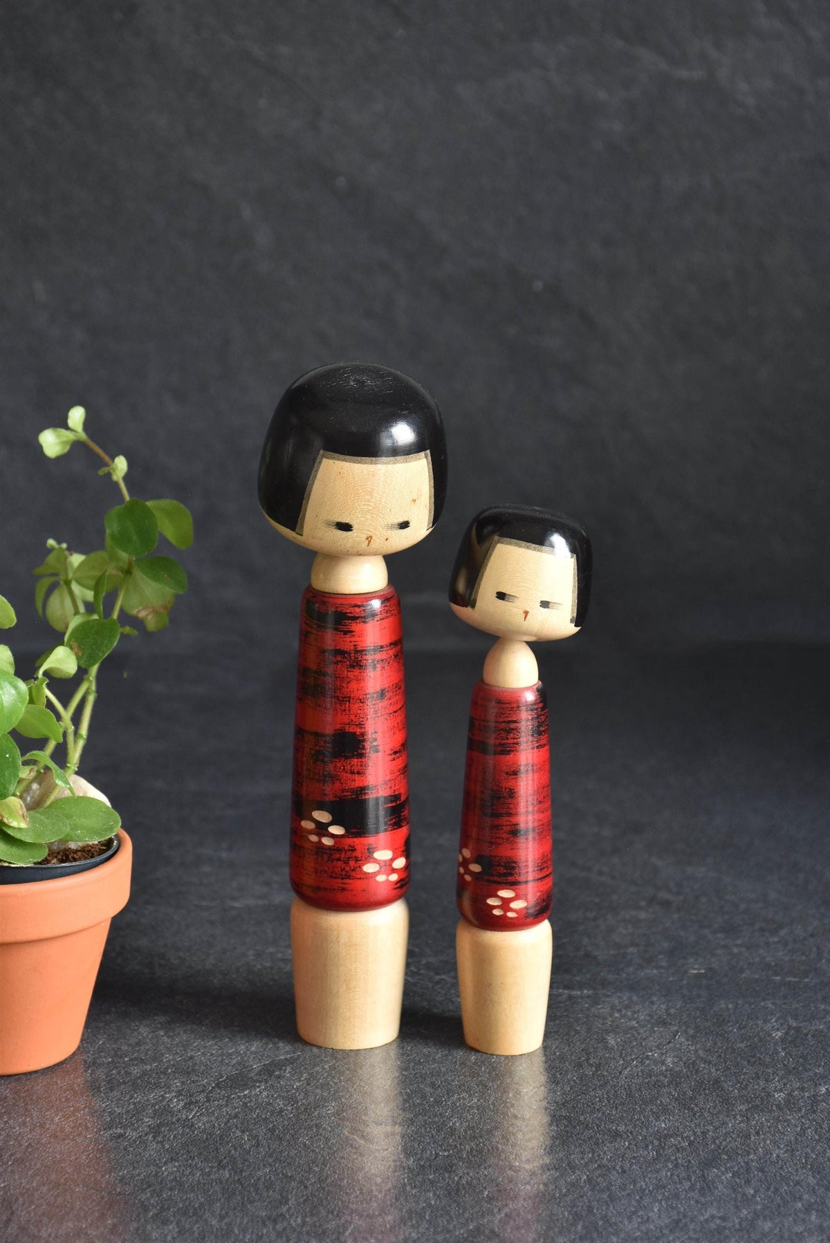 Aida Seihou Sosaku Kokeshi Sisters Red Bark Finish dolls standing side by side next to a small potted plant