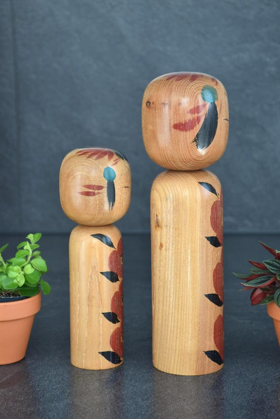 Takahashi Hashime kokeshi pair front view with potted green plants on a dark slate background