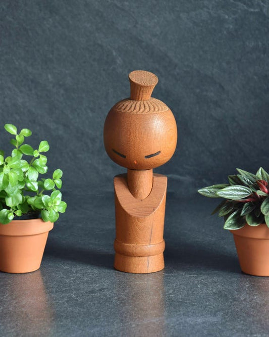 Aida Seihou Creative Kokeshi Doll standing upright with natural wood grain between two small potted plants