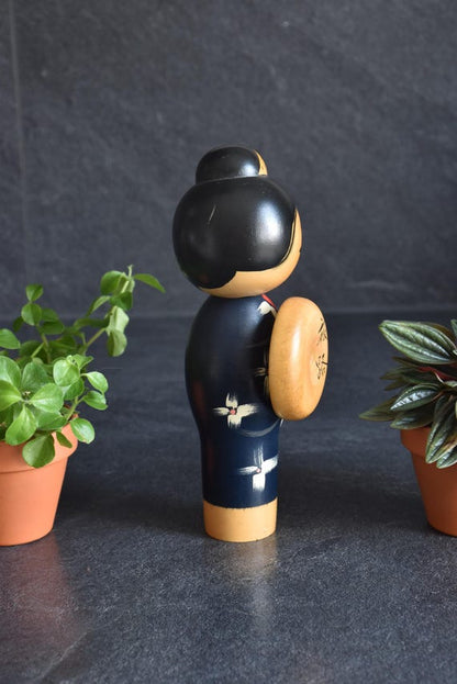Miyashita Hajime kokeshi doll shown from the side with its round wooden hat and potted plants