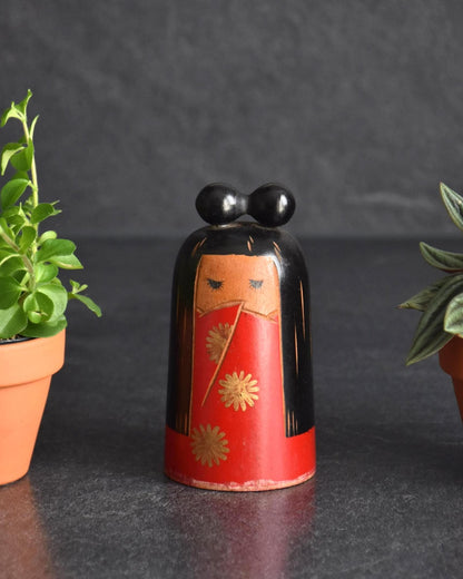 Japanese Kokeshi Doll With Black Ribbon displayed between two small potted plants on a dark surface