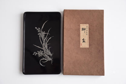 Black lacquer serving tray with silver orchid inlay shown vertically next to its textured storage box