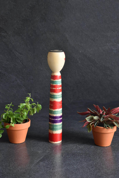 Tsuchiyu-Kei Kokeshi Doll Lathe-Spun Bands standing upright centered between potted plants against a dark surface