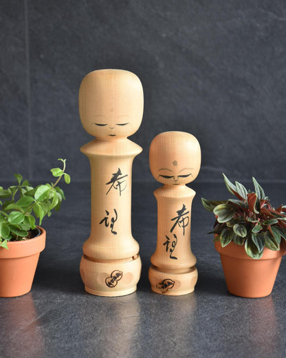 Isamu Ise Hiba Wood Kokeshi Dolls Hope Kanji pair displayed with potted plants.