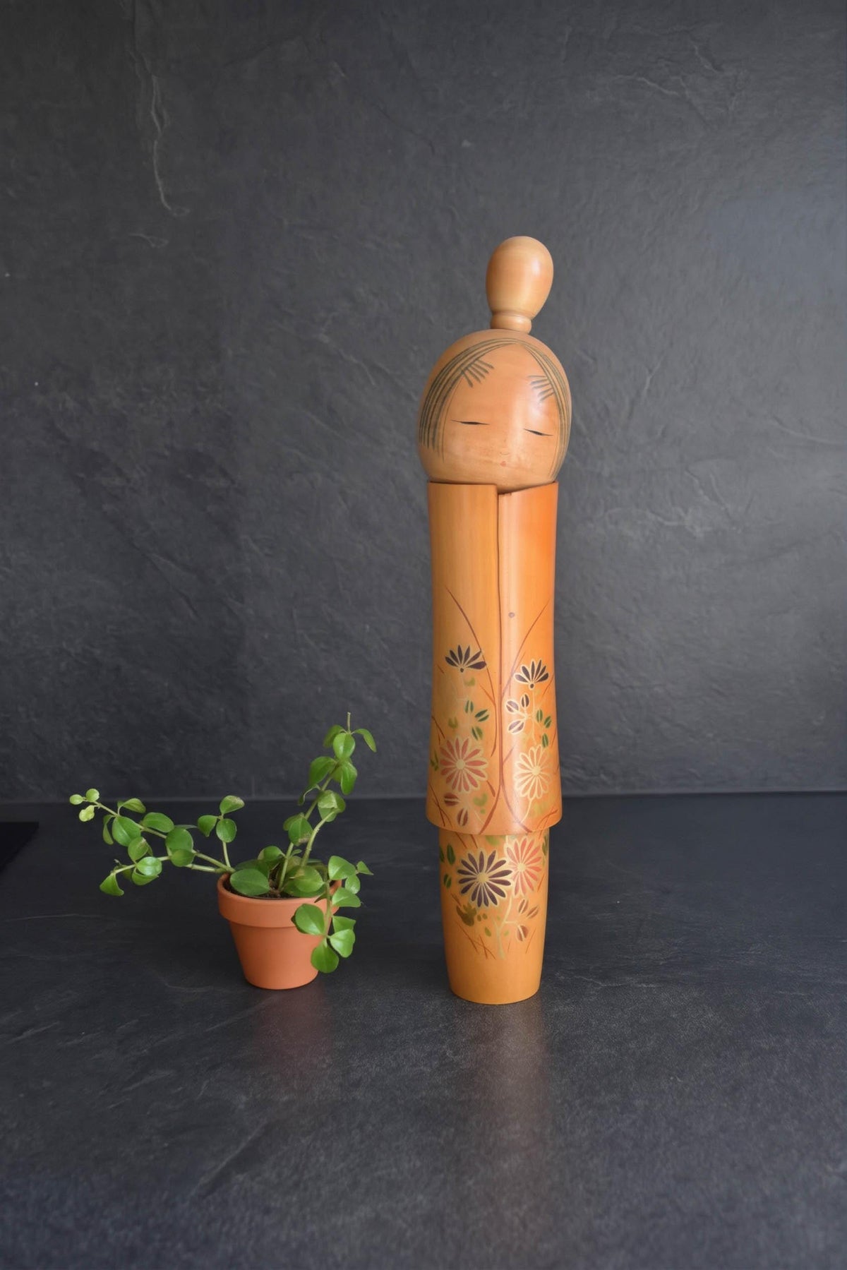 Sato Suigai Kiku No Omoi kokeshi doll standing upright next to a small potted green plant