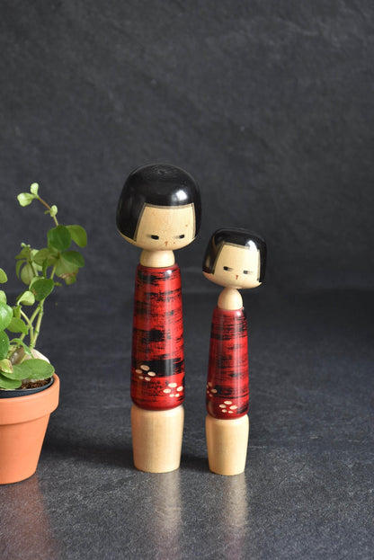 Aida Seihou Sosaku Kokeshi Sisters Red Bark Finish dolls standing side by side next to a small potted plant