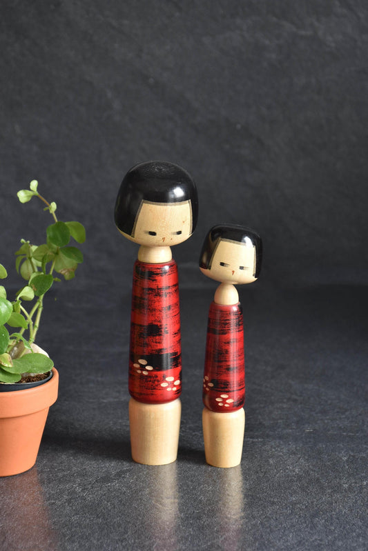 Aida Seihou Sosaku Kokeshi Sisters Red Bark Finish dolls standing side by side next to a small potted plant